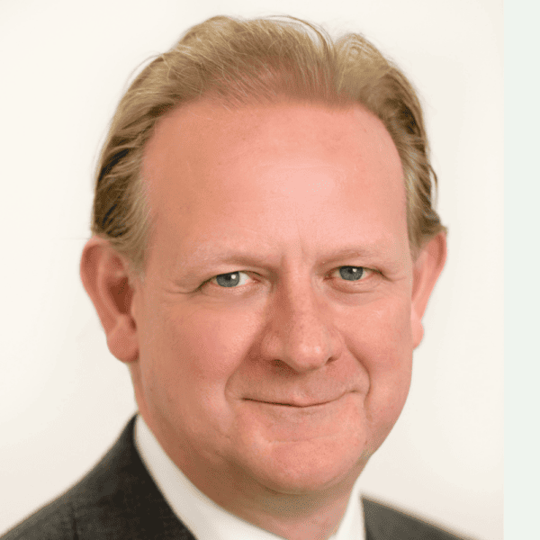 Headshot of Max Carter, from the shoulders up, smiling, in a charcoal blazer with short blonde hair