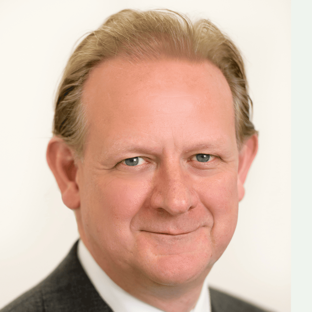 Headshot of Max Carter, from the shoulders up, smiling, in a charcoal blazer with short blonde hair