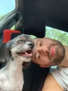 Image of a man and his white/black dog inside of a car smiling